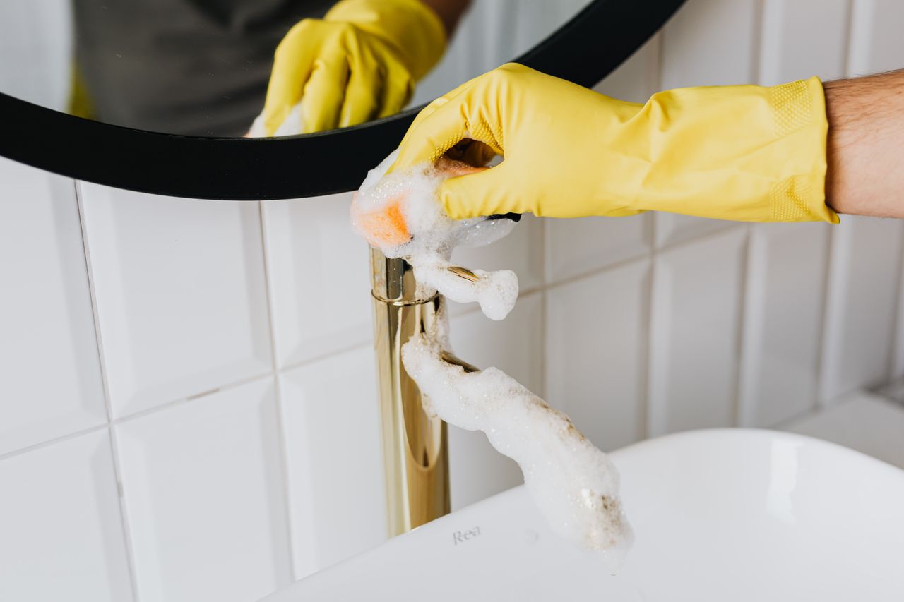 services-04 Personne anonyme portant des gants en latex jaunes qui nettoie un robinet doré avec une éponge dans une salle de bain moderne, pendant le ménage